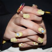 Hands with short, square-shaped press-on nails in yellow with white grid patterns, adorned with 3D flowers and stars.
