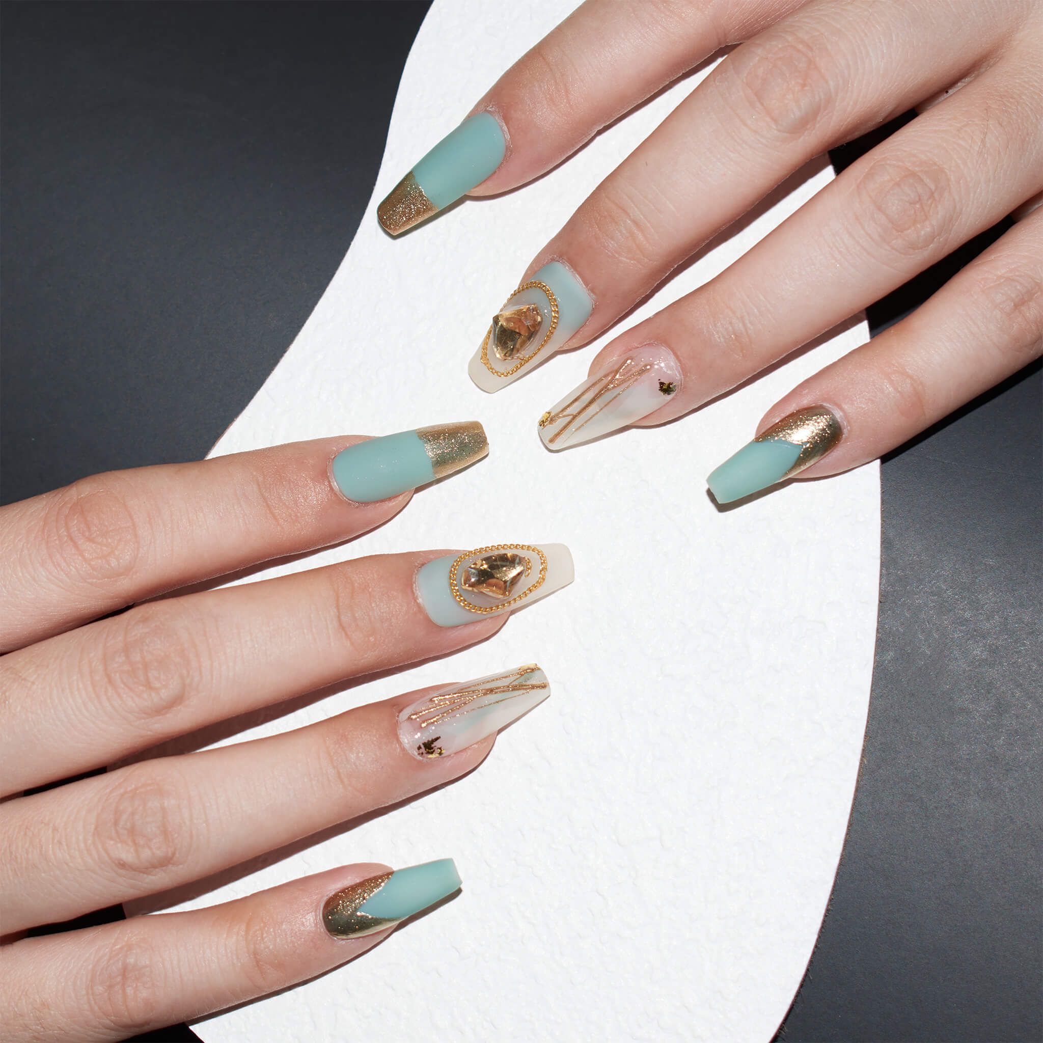 Hands with medium long, coffin-shaped press-on nails in matte aqua blue and glossy beige with gold accents, including glitter and geode patterns, against a black and white background.
