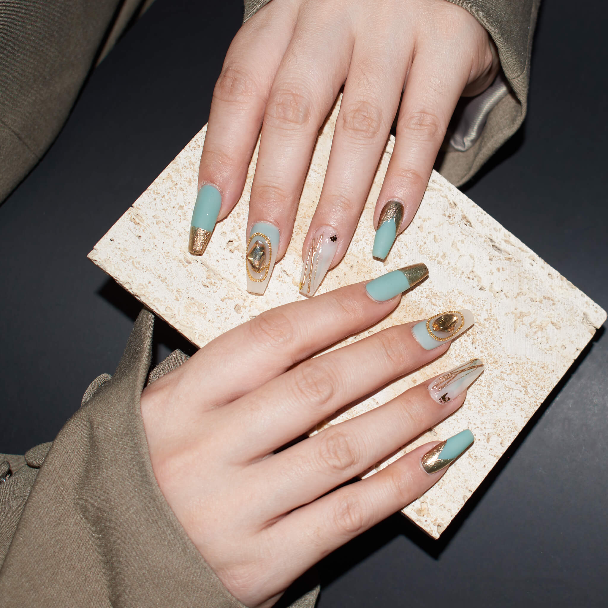 Hands with long, coffin-shaped press-on nails in matte aqua blue and glossy beige with gold accents, holding a textured beige object.