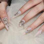 Elegant display of Monoschic Prismatic Princess Press-On Nails featuring dazzling rhinestones, pearls, and charms on a light pink nude base, arranged on a soft white fabric.