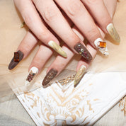 Hands with long, almond-shaped press-on nails in beige, brown, and taupe, adorned with marble patterns, gold accents, and 3D jewels.