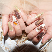 Close-up of hands with long, almond-shaped press-on nails in earthy tones, featuring gold accents, marble designs, and 3D gemstones.