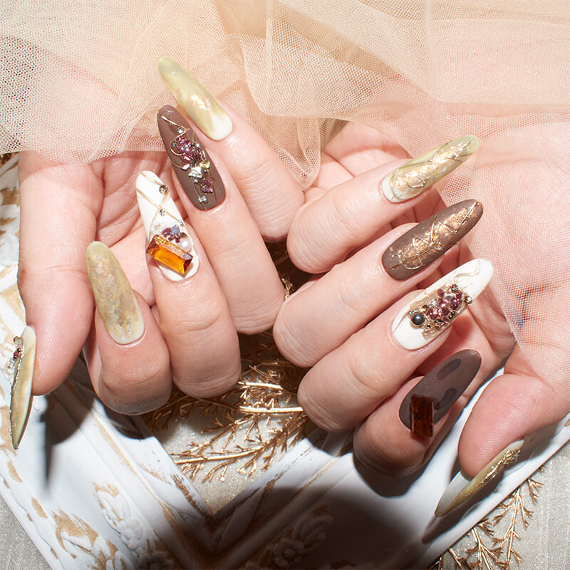 Close-up of hands with long, almond-shaped press-on nails in earthy tones, featuring gold accents, marble designs, and 3D gemstones.