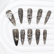stiletto-shaped press-on nails in dark, smoky hues with silver accents, including hearts, crosses, chains, and gems