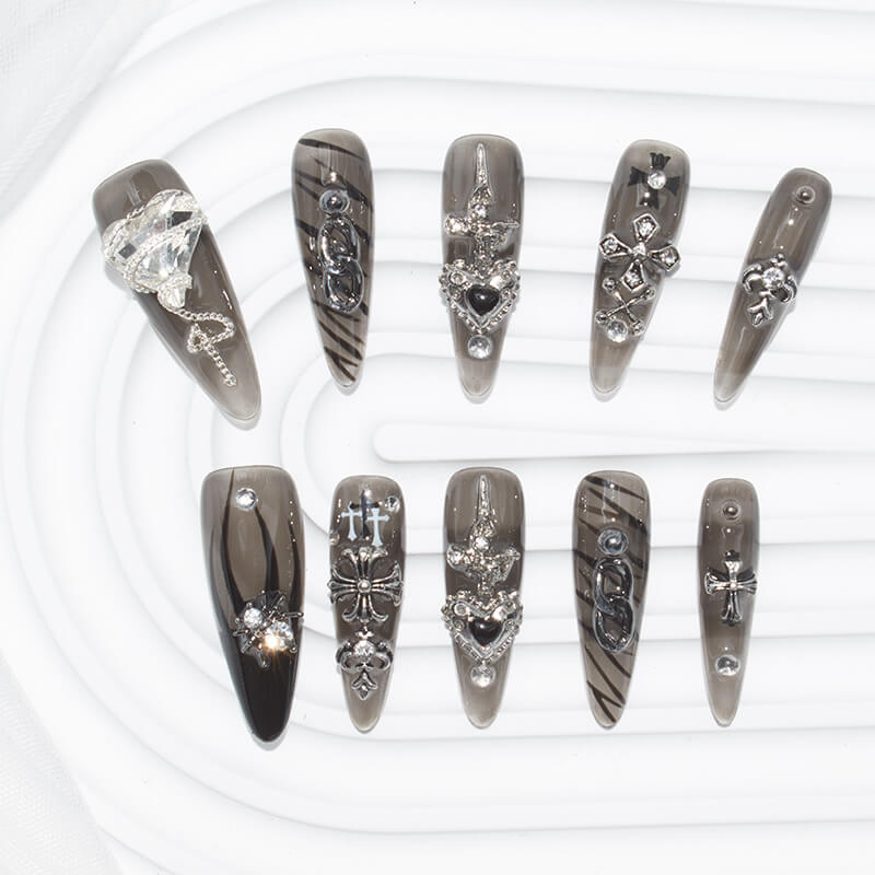 stiletto-shaped press-on nails in dark, smoky hues with silver accents, including hearts, crosses, chains, and gems